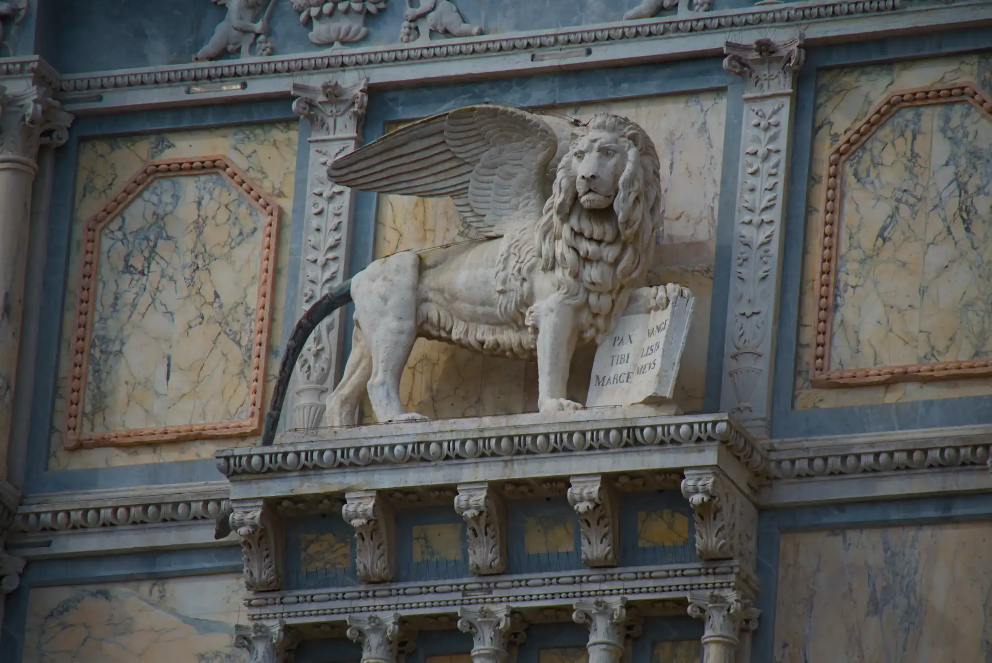 Sculpture en pierre d’un lion ailé tenant un livre ouvert, intégrée à une façade architecturale ornée de marbre et de motifs sculptés