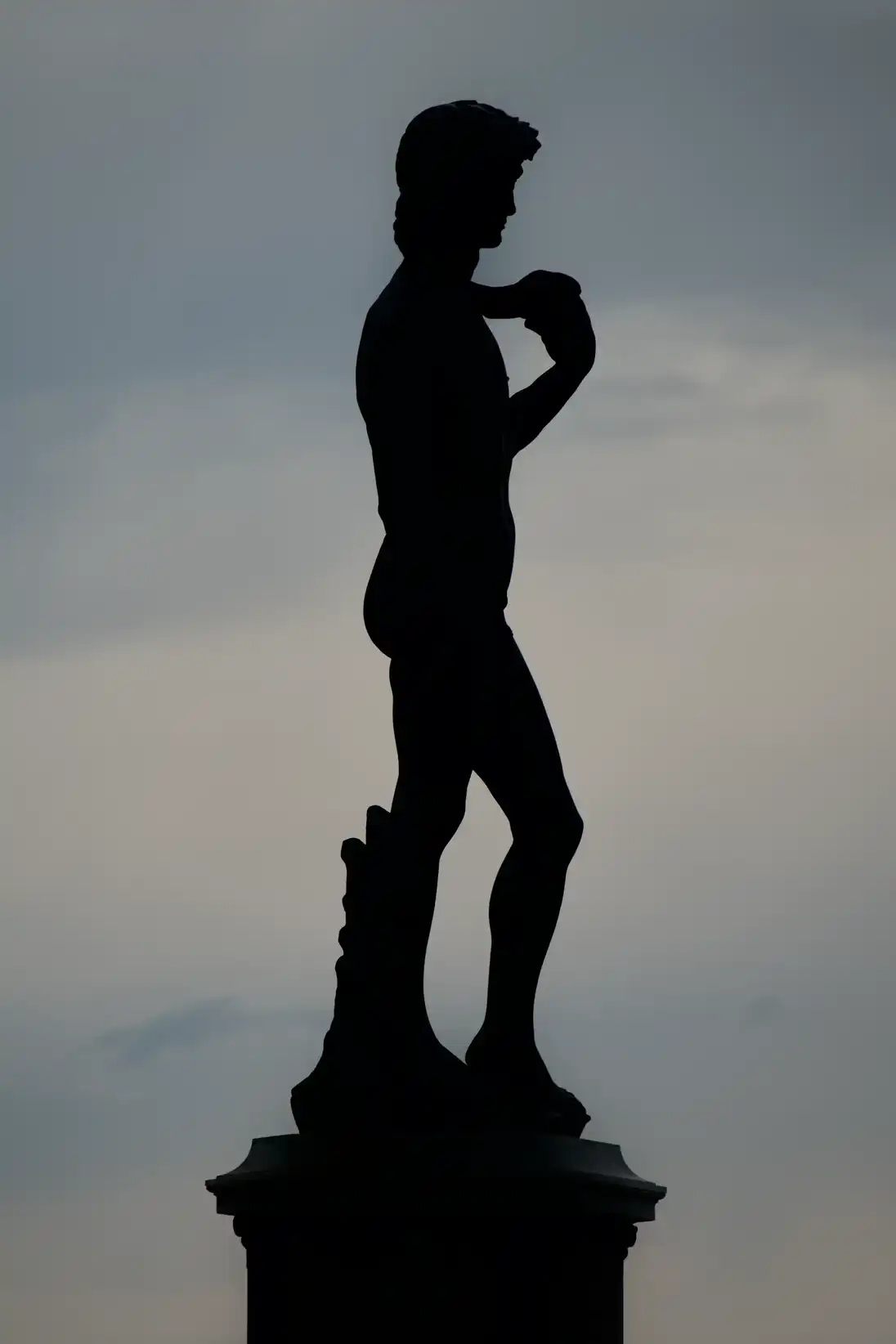 Silhouette sombre d’une statue masculine représentant un homme debout, le bras droit fléchi, sur un piédestal devant un ciel gris